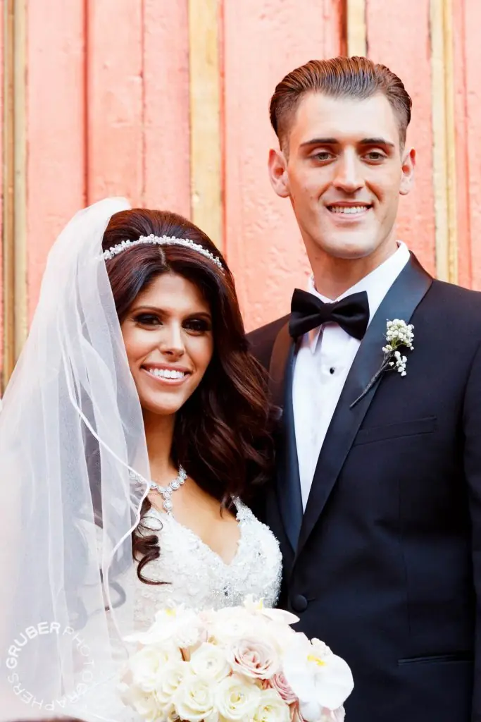 A bride in a white gown with a lace veil and a groom in a black tuxedo pose together in front of a pink background. The bride holds a bouquet of white and pink flowers.