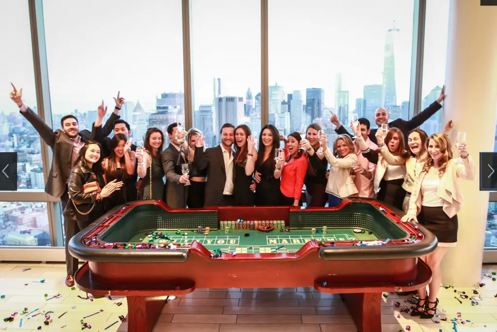 A large group of people stand around a casino craps table, cheering and celebrating. The background features tall buildings and a city skyline visible through large windows.